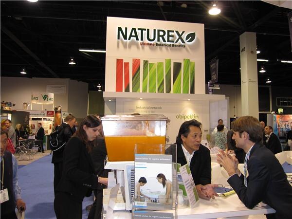 Two men talk at a counter under a Naturex sign with an obipektin banner, while a woman in dark clothing stands nearby next to a container of orange liquid.