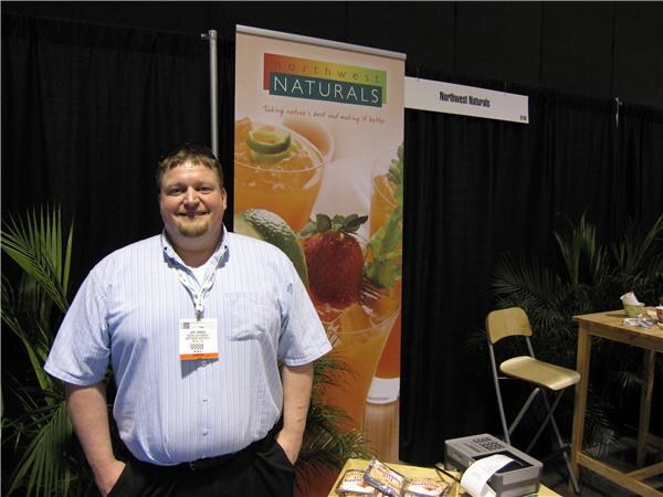A smiling man in a light blue striped shirt and name badge stands in front of a Northwest Naturals banner showing fruit and drinks, with product samples on a table.
