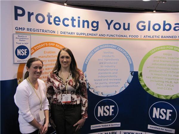 Two smiling women with lanyards stand in front of an NSF display reading Protecting You Globally, with panels on GMP registration and product certification.