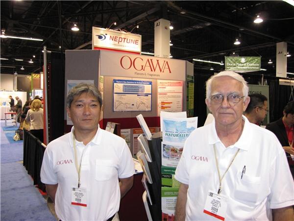 Two men in white Ogawa polo shirts with lanyards stand in front of the Ogawa Flavors & Fragrances booth, with a Neptune sign and a Draco Natural Products sign behind them.