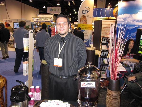 A man in a dark striped shirt and tie with a lanyard stands behind a counter with a dispenser labeled Organic Rooibos Superior, with EZ Brain, Gelmaxx and Pacific Rainbow signs behind him.
