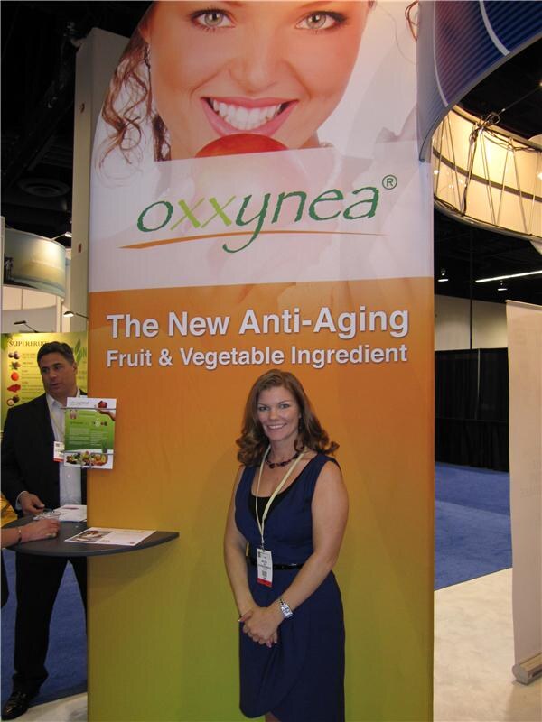 A woman in a navy dress and lanyard stands beside an oxxynea banner reading "The New Anti-Aging Fruit & Vegetable Ingredient"; a man in a dark suit stands behind her at a small table.