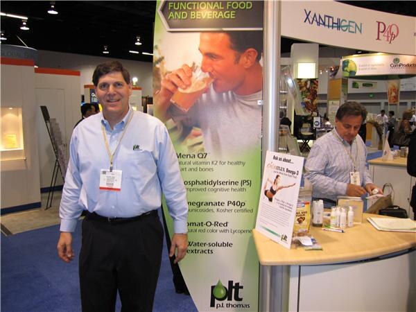 A man in a light blue shirt and lanyard stands by a P.L. Thomas banner for functional food and beverage ingredients, while another man sits at a counter below Xanthigen and P40p signs.