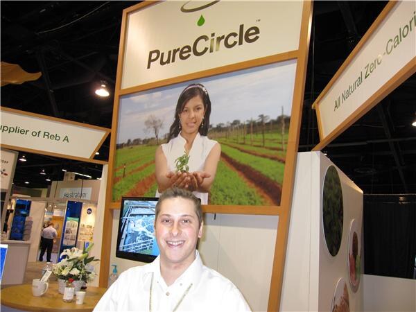 A smiling man in a white shirt sits at a table in front of a PureCircle sign showing a woman holding a green plant in a field, with a flower arrangement on the table.