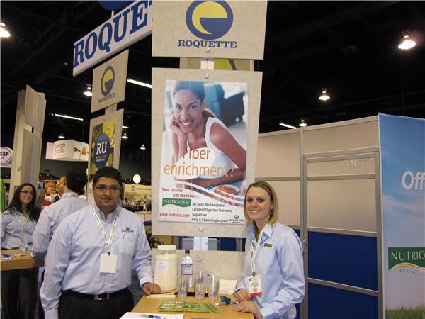 A man and a woman in light blue shirts and lanyards stand at a counter under Roquette signs and a Nutriose poster asking about fiber enrichment, with a container of powder on the table.