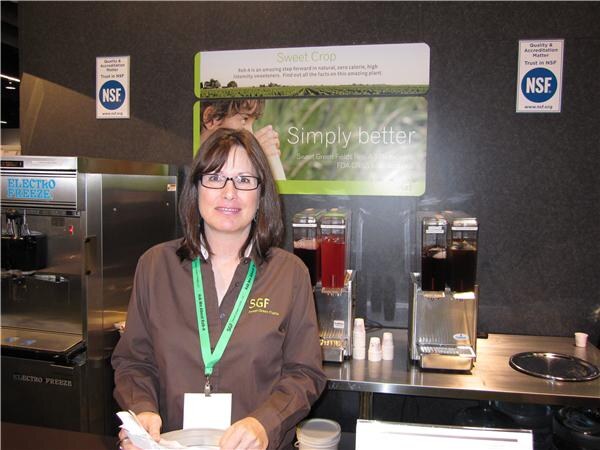 A woman with dark shoulder-length hair and glasses wears a brown SGF shirt and green lanyard at a booth with an Electro Freeze machine, beverage dispensers and a Simply better sign.
