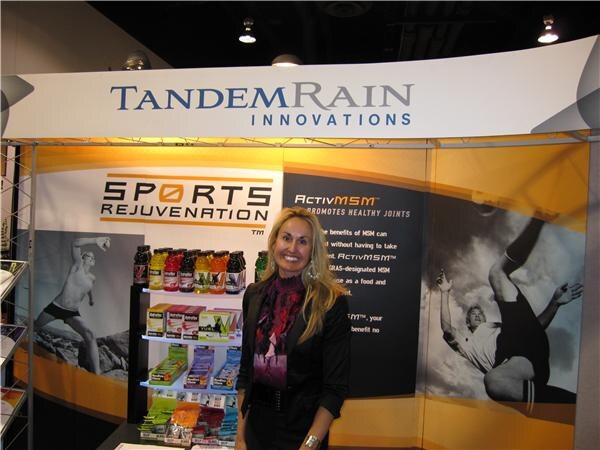 A woman with long blonde hair and a dark jacket stands in the Tandem Rain Innovations booth, with Sports Rejuvenation and ActivMSM graphics and shelves of bottled drinks and packaged products.