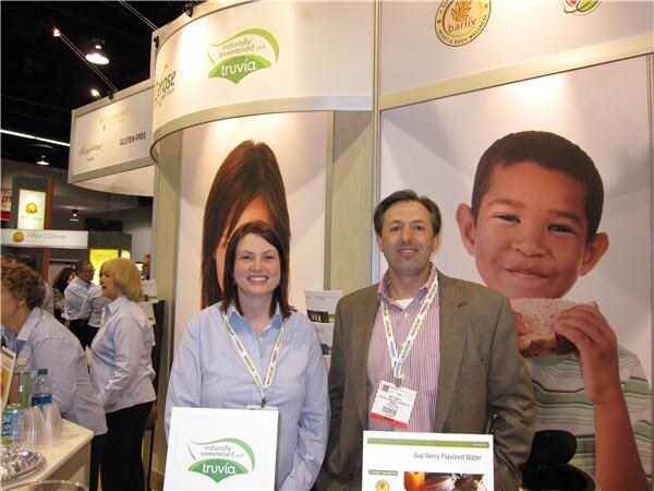 A woman in a light blue shirt and a man in a tan blazer, both wearing lanyards, stand in a booth with Truvia signs and a Barlív banner showing a smiling boy holding a cracker.