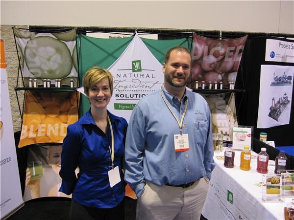 A smiling woman in a blue blouse and a bearded man in a light blue shirt stand in front of a Vi Natural Ingredient Solutions banner, with juice bottles on a table to the right.