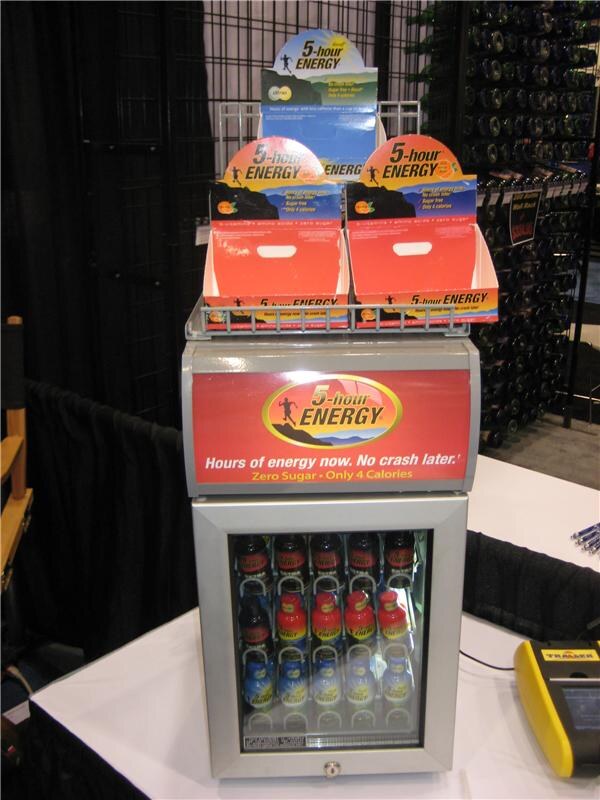 A small glass-door cooler stocked with 5-hour ENERGY bottles, topped with a red sign reading "Hours of energy now. No crash later." and cardboard display trays, on a draped table.