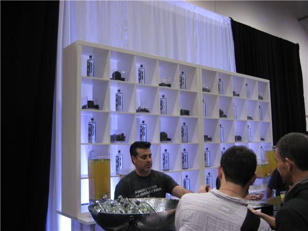A man in a dark T-shirt stands behind a white shelving unit holding 901 Silver Tequila bottles, with a bowl of bottles in ice and two people facing him in the foreground.