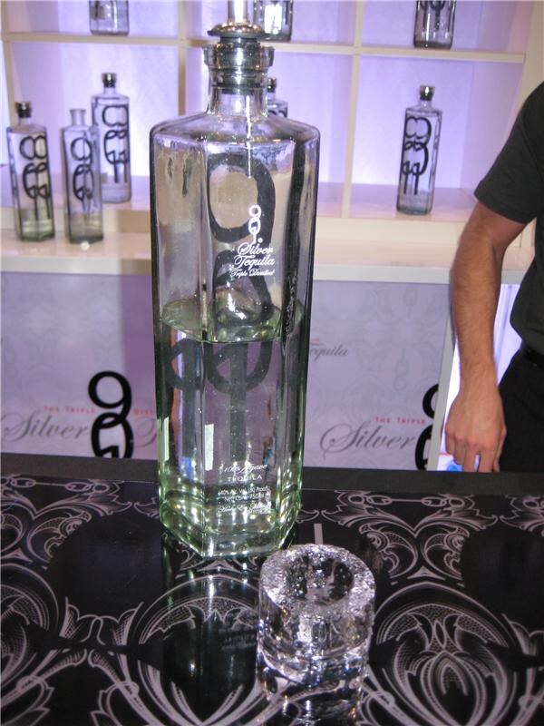 A large clear bottle of 901 Silver Tequila stands on a black patterned bar top beside an ice shot glass, with more bottles on lit shelves behind and a person's arm at right.
