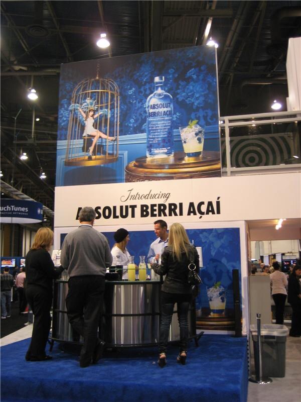 A two-level booth with an Introducing Absolut Berri Açaí banner and a bottle graphic, with staff behind a round bar on a blue carpeted stage and visitors standing in front.