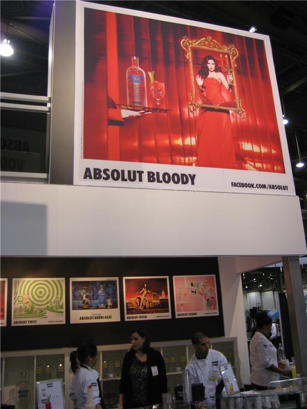 A large Absolut Bloody advertisement with a woman in a red gown hangs above the booth, with smaller Absolut posters and staff in white jackets below.