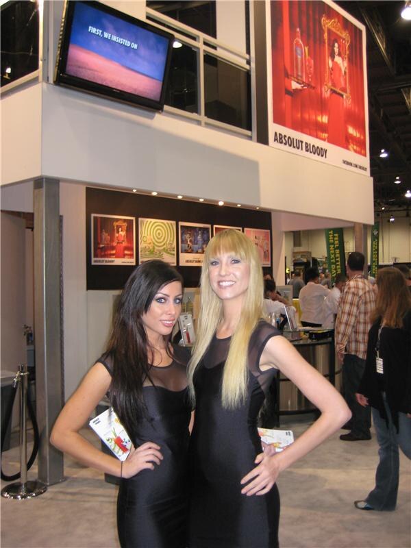 Two women in black dresses, one with long dark hair and one with long blonde hair, pose in front of the Absolut booth with an Absolut Bloody banner and posters behind them.