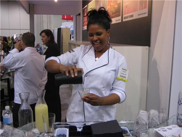 A smiling woman in a white chef's jacket pours from a black shaker into a small cup at the Absolut booth, with a pitcher of yellow juice and other attendees behind her.