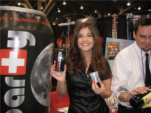 A smiling woman in a black top holds a can of Afterparty and a clear cup beside a giant inflatable can, while a man in a white shirt and black tie pours from a bottle at right.