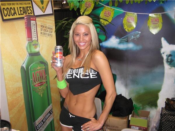 A smiling blonde woman holds a can in front of Agwa de Bolivia displays, including a large green bottle graphic, coca leaves signage and a llama backdrop.