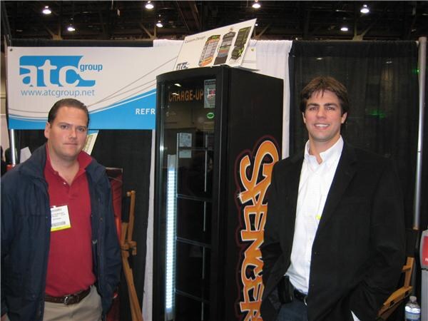 Two men stand in an atc group booth beside a tall black Spenger-branded cooler. One wears a red polo and a show badge; the other wears a dark blazer over a white shirt.
