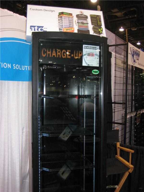 A tall black glass-door cooler labeled Charge-Up with empty wire shelves, under an atc group Custom Design sign showing several cooler models.