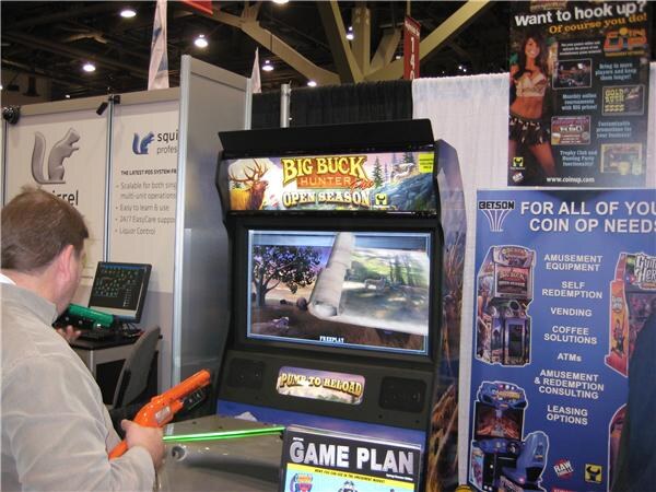 A man in a gray sweater aims an orange plastic gun at a Big Buck Hunter Pro Open Season arcade cabinet. A Betson banner and a Squirrel sign are visible beside it.