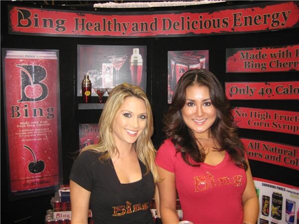 Two smiling women in Bing-branded shirts stand in front of a red and black Bing display. Signs read "Healthy and Delicious Energy" and show a Bing can with a cherry logo.