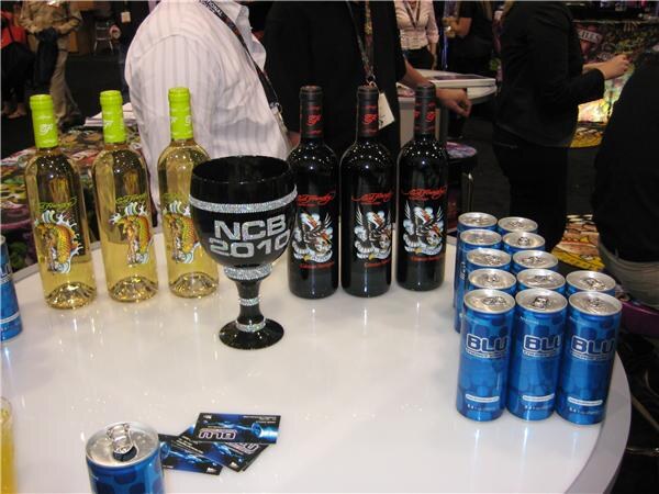 White round table displaying three bottles of white wine and three black bottles with Ed Hardy labels, a black glitter goblet reading NCB 2010, and several blue Blu energy drink cans.