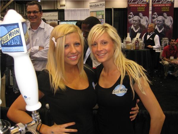 Two smiling blonde women in black Blue Moon tops stand beside a Blue Moon tap handle. MicroSale POS and Arnold Palmer Half & Half Lemonade signs are in the background.