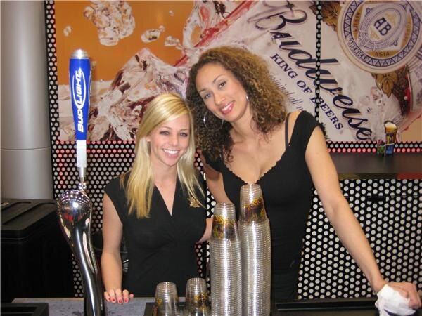Two smiling women in black tops stand behind a bar with stacked plastic cups. A Bud Light tap handle is at left and a Budweiser graphic is on the wall behind them.