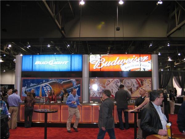 A trade-show booth with a long lit bar counter under illuminated Bud Light and Budweiser signs. Attendees stand and walk in front of it on red carpet in a large exhibition hall.