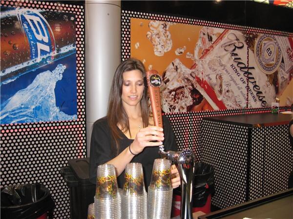 A woman with long brown hair pulls a draft tap handle at a bar. Stacks of plastic cups sit in front of her, with Bud Light and Budweiser graphics on the wall behind.