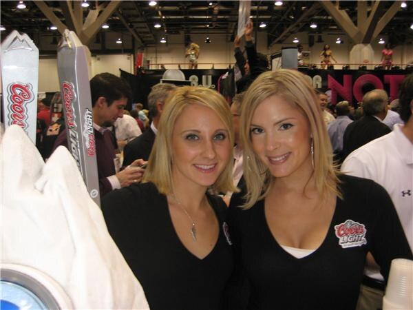 Two smiling blonde women in black tops, one with a Coors Light logo, stand beside Coors Light tap handles on a busy show floor with attendees behind them.