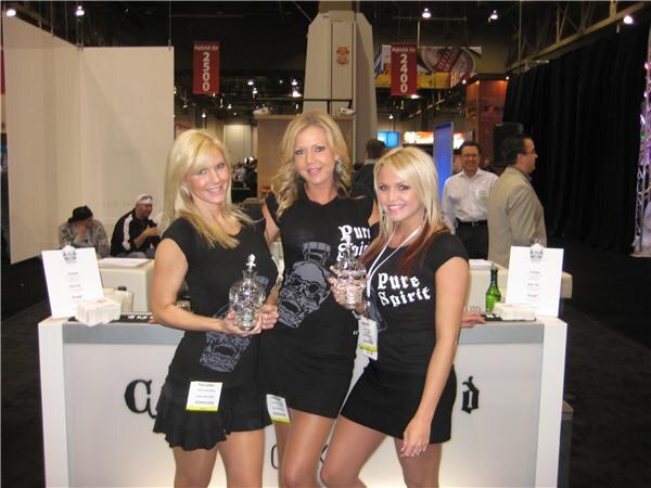 Three women in black Pure Spirit skull T-shirts stand behind a white counter, two holding clear skull-shaped Crystal Head bottles, with other attendees in the background.