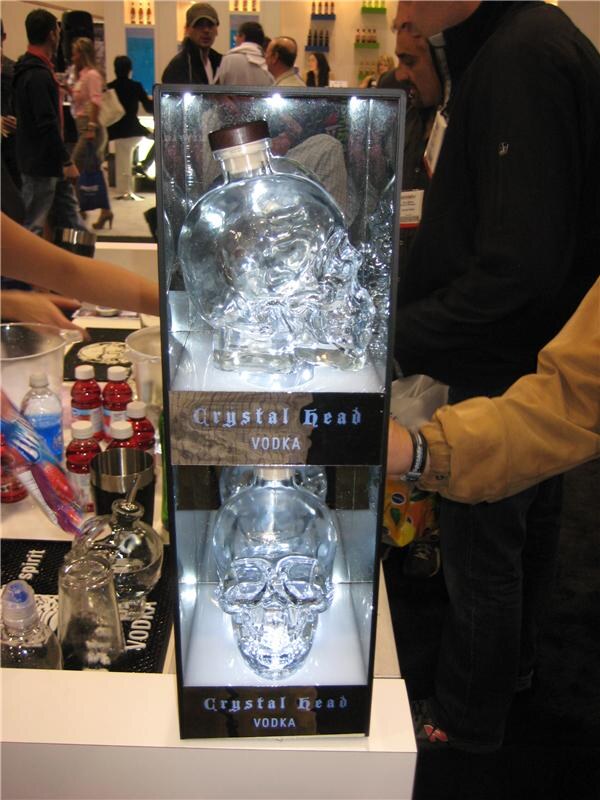 Lit display case holding two clear skull-shaped Crystal Head Vodka bottles, with attendees standing nearby and bar supplies on a counter at left.