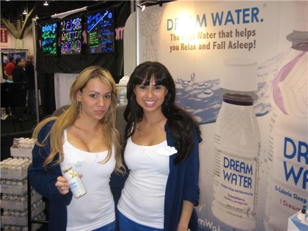 Two women in white tops and blue cardigans stand before a Dream Water banner, one holding a small bottle, with product cases and lit message boards behind them.