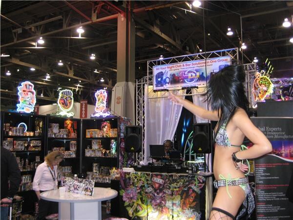 Booth with neon Ed Hardy signs, lit display shelves, a DJ table with tattoo-art graphics, and a dancer in a bikini and fishnets in the foreground.