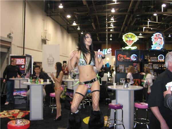 A dancer in a patterned bikini and fishnets stands among high tables and stools near neon Ed Hardy signs, with a Marvel banner and attendees behind her.