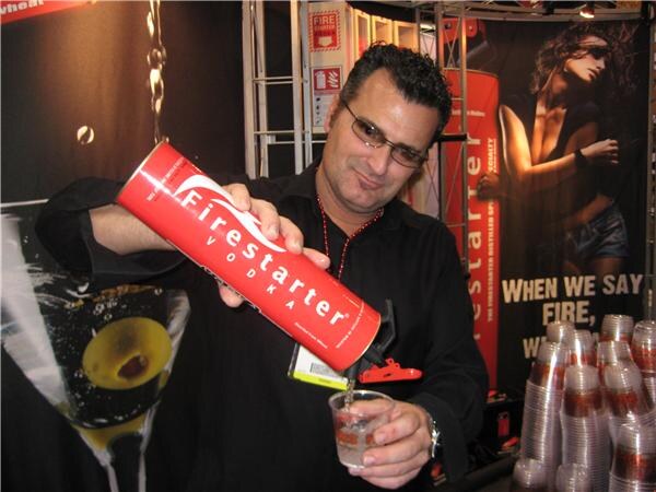 A man with glasses and a black shirt pours a clear drink from a red Firestarter Vodka canister into a small plastic cup, with Firestarter posters and stacked cups behind him.