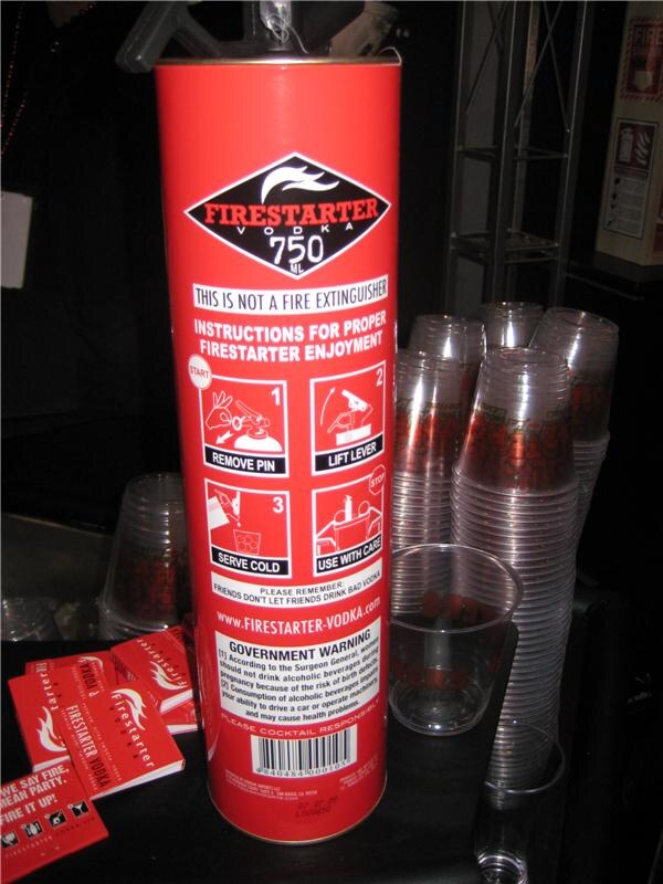 Back of a red tube-shaped Firestarter Vodka 750 ml package with instructions, next to a stack of clear plastic cups and red matchbook-style cards on a dark table.
