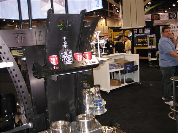 A black metal Fry Fabrications bar display holds a Jack Daniel's bottle and three Coca-Cola cans on a shelf labeled Jack & Coke, with other booths and attendees in the background.