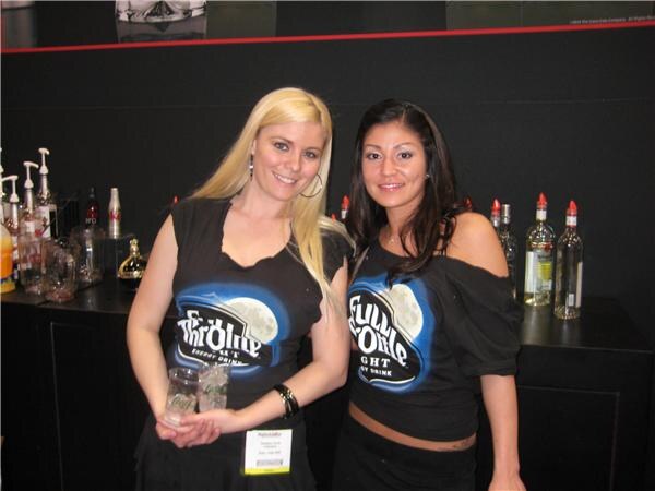 Two smiling women in black Full Throttle Night energy drink shirts stand at a dark bar, one holding two small glasses, with liquor bottles and syrup pumps behind them.