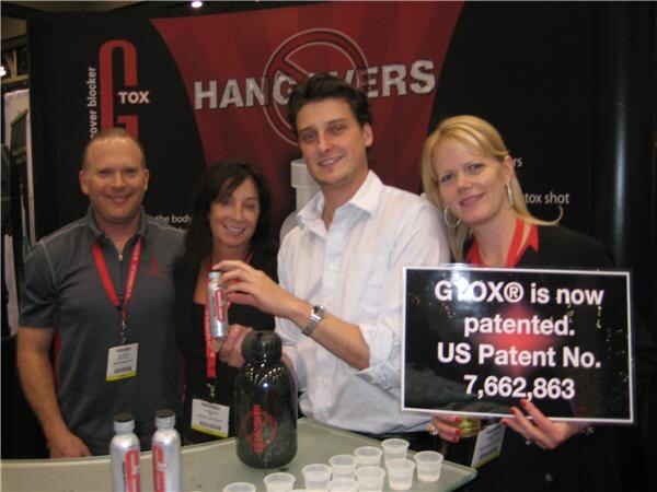 Four people smile behind a G-Tox counter with small cups and bottles; a woman holds a sign reading G-Tox is now patented, US Patent No. 7,662,863, and a man holds a G-Tox can.