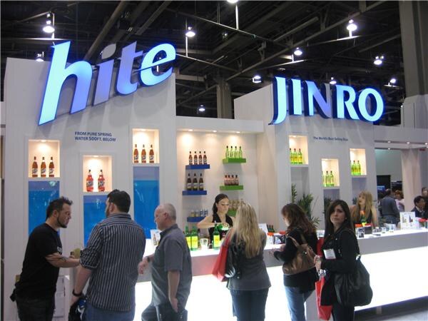 A white booth with lit Hite and Jinro signs, shelves of bottles and a long counter where several visitors stand and a woman works behind it.