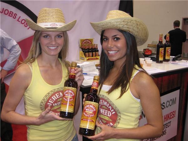 Two smiling women in straw cowboy hats and yellow Jeremiah Weed tank tops each hold a bottle of Jeremiah Weed Sweet Tea at a booth.