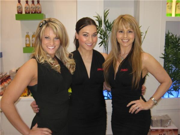 Three smiling women in black dresses pose together in the Jinro booth, with shelves of bottled drinks and green plants behind them.