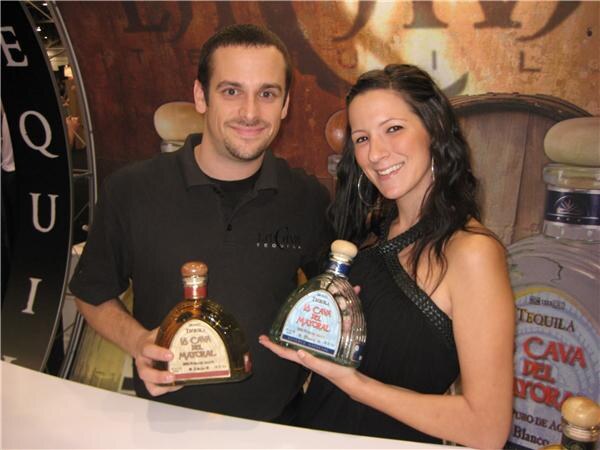 A smiling man in a black polo and a smiling woman in a black dress each hold a bottle of La Cava del Mayoral tequila at a booth with a large bottle and barrel graphics behind them.