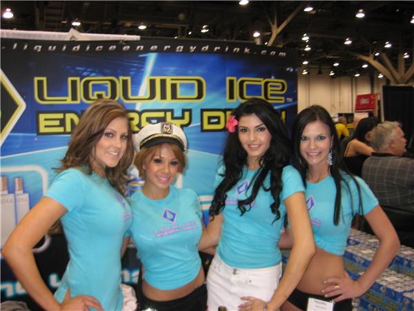 Four smiling women in light blue Liquid Ice shirts, one in a white captain's hat, pose in front of a Liquid Ice Energy Drink banner, with stacked cases of drinks at right.