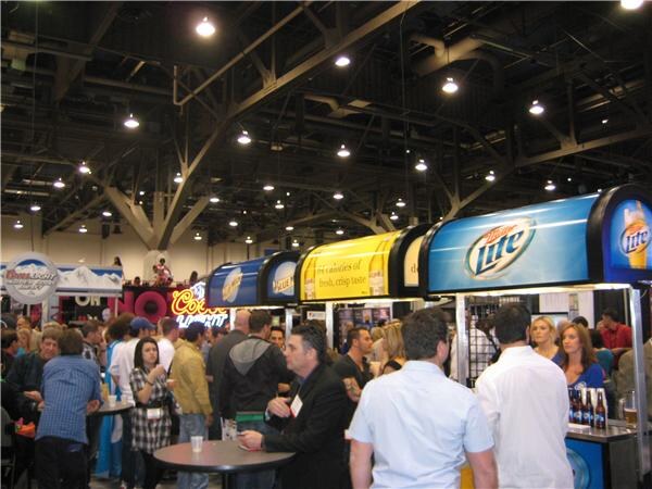 Attendees gather around lit bar-style booths with Miller Lite and Coors signs under the exposed rafters and lights of a convention hall.