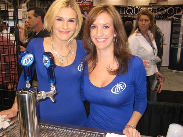 Two smiling women in blue Miller Lite shirts stand behind a double tap with Miller Lite handles. A Liquor Booty sign and other attendees are in the background.