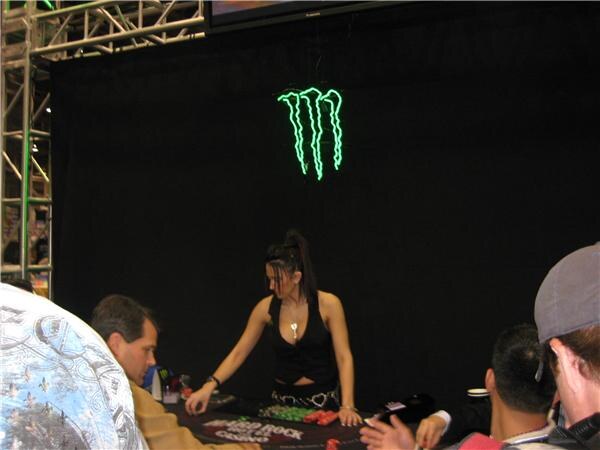A woman deals cards at a Hard Rock poker table beneath a green neon Monster logo while several seated players are seen from behind and the side.